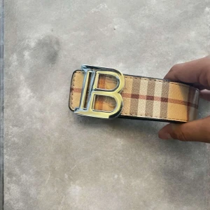 Burberry rutiga skärp med B-spänne - Två breda Burberry-bälten med rutigt mönster. B-logga som spänne. Ett bälte i beige, det andra i svart och grått. Båda i skinn med Burberry-vibe, perfekt för känna sig lyxig. Den passar dig me alla stilar 