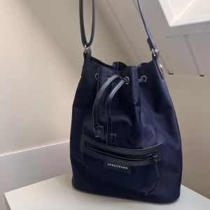 Mörkblå axelväska från Longchamp - Snygg mörkblå axelväska från Longchamp i klassisk bucket-modell. Väskan har justerbar axelrem, dragsko upptill och en praktisk dragkedjeficka framtill. Tillverkad i slitstark syntet med detaljer i skinn. Perfekt för dig som vill ha en stilren och rymlig väska.