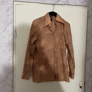 Mönstrad skjorta från Beat, stl 40 - Retro skjorta från Beat i beige med små prickiga och grafiska mönster i brunt. Skjortan har klassisk krage, långa ärmar och knäppning framtill. Tillverkad i en mix av polyester och bomull, perfekt för dig som gillar vintagevibbar och unika prints.