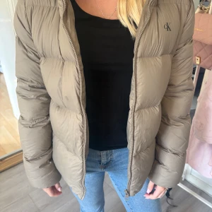 Beige pufferjacka från Calvin Klein - Säljer en beige pufferjacka från Calvin Klein Jeans med diskret CK-logga på bröstet. Jackan har en klassisk pufferlook med breda kanaler, dragkedja framtill och lång ärm. Perfekt för kyliga dagar och riktigt snygg till jeans eller mjukis.