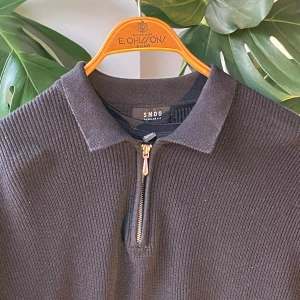 Svart stickad tröja med halv dragkedja - Svart stickad tröja från SMOG med klassisk krage och halv dragkedja framtill. Tröjan har ribbad struktur och lång ärm, perfekt för lager-på-lager under kyliga dagar. Enkel och stilren design som funkar till det mesta.