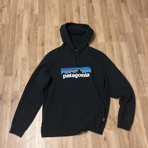 Svart Patagonia hoodie med logga - Svart hoodie från Patagonia i storlek M med klassisk logga framtill i blått, rött och vitt. Tröjan har huva med snörning och en stor magficka. Tillverkad i bomull och polyesterblandning, perfekt för chill dagar eller när du vill ha en clean streetstyle.