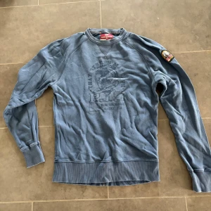 Blå sweatshirt från Parajumpers - Snygg blå sweatshirt från Parajumpers med tryck framtill och patch på ena ärmen. Tröjan har rund halsringning, långa ärmar och ribbade muddar vid ärmslut och nederkant. Perfekt för en avslappnad och cool stil.