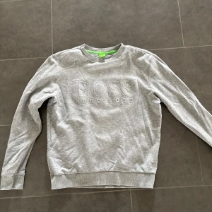 Grå sweatshirt från Hugo Boss - Säljer en stilren grå sweatshirt från Hugo Boss med broderad logga framtill. Tröjan har rund halsringning, långa ärmar och ribbade muddar. Perfekt för en chill och snygg look.