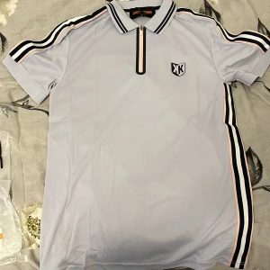 Ljusblå polo från  Foot korner med dragkedja - Snygg grå, vit och rosa på sidan polo från Foot korner med korta ärmar, krage och dragkedja framtill. T-shirten har svarta och rosa ränder längs axlar och sidor samt ett broderat märke på bröstet. Perfekt för en sportig och trendig look. Den är helt ny och har kvar etiketten!