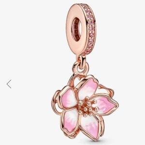 Pandora blomcharm i roséguld - Superfin charm från Pandora i roséguld med en hängande blomma i rosa och vitt emalj. Ringen är dekorerad med små rosa stenar som glittrar. Perfekt detalj till ditt armband om du vill ha en söt och färgglad look. Orginal pris 1000,- sek