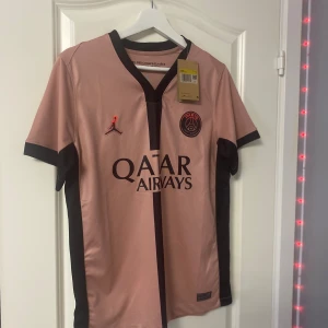 PSG x Jordan rosa fotbollströja - Snygg PSG fotbollströja i samarbete med Jordan. Tröjan är rosa med svarta detaljer, svart vertikal rand och svarta muddar. Broderad PSG-logga och Jumpman-logga på bröstet. Tillverkad i lätt och ventilerande Dri-FIT-material. Perfekt för dig som älskar fotboll och streetstyle.