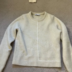Beige stickad tröja från Zara - Supermjuk beige stickad tröja från Zara med rund hals och lång ärm. Tröjan har en clean look med en vit dekorativ söm som går både fram och bak. Använd fåtal gånger.
