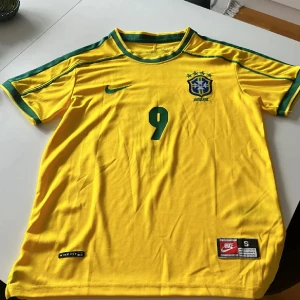 Brasilien Ronaldo 9 fotbollströja Nike  - Gul Brasilien fotbollströja från Nike med gröna detaljer, nummer 9 och Ronaldo på ryggen. Klassisk design med korta ärmar, rund hals och broderat landslagsemblem på bröstet. Tillverkad i lätt och ventilerande material, perfekt för fotbollsälskare.