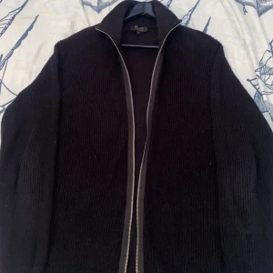 Svart stickad kofta från INDICODE - Svart stickad kofta från INDICODE med hel dragkedja framtill och hög krage. Tröjan har ribbad struktur och långa ärmar, perfekt för lager-på-lager-stil. Snygg detalj med liten metallbricka nertill. Passar dig som gillar enkel och clean look. Aldrig använt passar inte mig. Eftersom den är för stor.