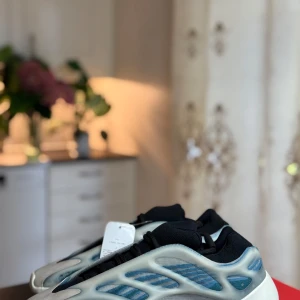 Adidas Yeezy 700 V3 blå sneakers - Säljer ett par Adidas Yeezy 700 V3 sneakers i blått och vitt med svarta detaljer. Skorna har en futuristisk design med mesh och syntetmaterial, samt snörning och rund tå. Den tjocka vita sulan ger en cool siluett och de blå inslagen gör dem riktigt unika.