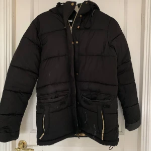 Svart pufferjacka från Jeane Blush XS - Svart pufferjacka från Jeane Blush i storlek XS. Jackan har huva, guldiga dragkedjor och knappar, samt stora fickor framtill. Insidan är beige och jackan är vadderad för extra värme. Perfekt för kalla dagar och har en clean, enkel look.