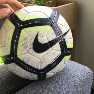 Nike Strike fotboll vit/svart/gul - Säljer en Nike Strike fotboll med Aerowtrac-teknologi. Bollen är vit med svarta och neongula detaljer samt tunna orange linjer. Tillverkad i slitstarkt syntetmaterial med texturerad yta för bättre grepp och kontroll. Perfekt för matcher och träning.