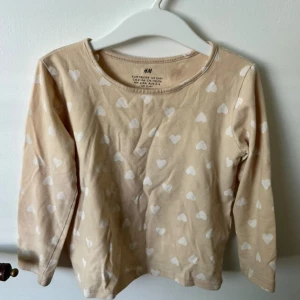 Beige långärmad tröja med hjärtan H&M - Supersöt beige långärmad tröja från H&M med vita hjärtan över hela. Mjuk och skön i bomull, perfekt för dig som gillar gulliga prints. Rund halsringning och normal passform, passar till både jeans och kjol.
