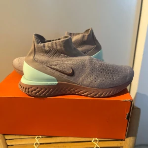 Nike Epic React Flyknit sneakers - Nike Epic React Flyknit sneakers i ljusgrått med stickad ovandel och elastisk strumpliknande passform. Sulan är vågigt mönstrad i brunt och ljusblått. Perfekta för dig som gillar stilrena och bekväma skor med modern design. Modellen finns ej att få tag på ! Utgående modell