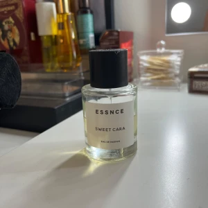 ESSNCE Sweet Cara Eau de Parfum - Fräsch parfym från ESSNCE med namnet Sweet Cara. Kommer i en stilren, rund glasflaska med svart lock och enkel vit etikett. Parfymen har en ljusgul ton och känns modern och minimalistisk i designen. Perfekt för dig som gillar enkla men snygga detaljer.