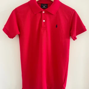 Röd pikétröja från Polo Sport Ralph Lauren - Klassisk röd pikétröja från Polo Sport Ralph Lauren i 100% bomull. Tröjan har korta ärmar, krage och två knappar framtill. Broderad marinblå logga på bröstet ger en sportig och stilren vibe. Perfekt för dig som gillar preppy och tidlös stil.