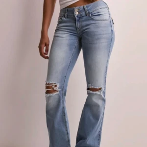 Ljusblå bootcut jeans med slitningar - Snygga ljusblå jeans med bootcut passform och slitna hål över knäna. Jeansen har dubbla knappar i midjan, klassiska fickor fram och bakfickor med lock och knapp. Perfekta för en avslappnad och trendig look.