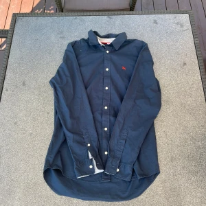 Mörkblå skjorta med röd logga - Snygg mörkblå skjorta med klassisk krage och vita knappar. På bröstet finns en liten röd broderad logga som ger en stilren touch. Skjortan har lång ärm och är tillverkad i mjuk bomull, perfekt för en clean och avslappnad look.