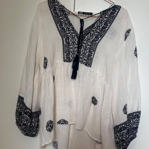 Vit blus med broderi från Zara - Luftig vit blus från Zara med mörkblå broderade detaljer och knytning med tofs framtill. Blusen har v-ringning, långa ärmar med broderade muddar och ett bohemiskt mönster över hela plagget. Perfekt för en chill och stilren look.