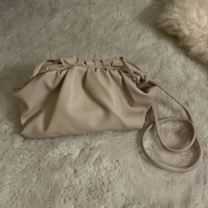 Beige clutch väska med snörning - Säljer en superfin beige axelväska i mjukt syntetmaterial. Väskan har en cool rynkad design upptill med snörningsdetalj och lång axelrem. Perfekt storlek för det viktigaste och stilren look som passar till många outfits.