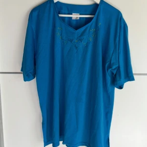 Blå blus med broderi och v-ringning - Säljer en blå blus med v-ringning och broderade blommor vid halsen. Toppen har korta ärmar och är gjord i mjukt bomullstyg, perfekt för en chill och stilren look. Passar dig som gillar enkla men snygga detaljer.