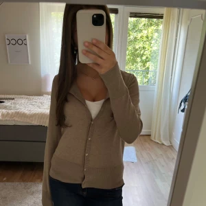 Beige kofta från Holly & White - Snygg beige kofta från Holly & White med rund halsringning och knappar framtill. Långärmad och i mjukt, tunt material som passar perfekt till jeans eller kjol. Enkel och stilren design som är lätt att matcha.