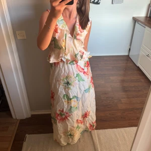 Blommig långklänning med volang - Säljer denna heöt nya långklänningen med prislapp kvar!  Den har ljus bas med stora blommor i rött, grönt och gult. Klänningen har v-ringning och volangdetaljer framtill och bak. Knytning i nacken för extra detalj. Perfekt för sommarens alla tillfällen! Klänningen är verkligen så vacker!!🌸