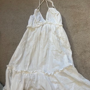 Vit långklänning från Divided XS - Superfin vit långklänning från Divided i storlek XS. Klänningen har smala axelband som knyts i nacken, v-ringad urringning och volangdetaljer. Den är luftig och har en romantisk känsla, perfekt för varma dagar. Aldrig använd.