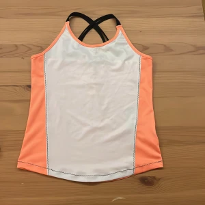 Sportig linne från Blue Seven, strl 152 - Snyggt sportlinne från Blue Seven i vitt och persikoorange med svarta korsade axelband. Perfekt för träning eller aktiva dagar. Tillverkat i ett mjukt och stretchigt syntetmaterial som andas. Snygga kontrastsömmar längs sidorna ger en cool look.