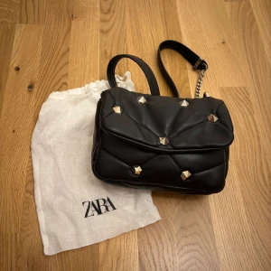 Svart väska från Zara - Snygg svart axelväska från Zara med quiltad design och coola guldiga nitar. Väskan har kedjedetalj på axelremmen och kommer med original dustbag.💓