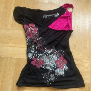 Svart blommig topp från Jennifer Taylor - Storlek L men passar S mer Unik svart topp med blommigt mönster i rosa och silver från Jennifer Taylor. Ena axeln har en cool ringdetalj och rosa tyg, medan den andra har en kort puffärm. Toppen har v-ringning och är gjord i mjuk bomull, perfekt för dig som gillar att sticka ut.