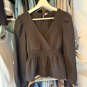 Svart omlottopp från H&M i XS - Snygg svart omlottopp från Divided i storlek XS. Toppen har v-ringning, långa ärmar och smockad midja som ger en fin form. Perfekt att styla med jeans eller kjol för en trendig look. 