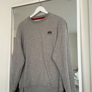 Grå sweatshirt Alpha Industries M - Snygg grå sweatshirt från Alpha Industries med svart logga på bröstet. Tröjan har rund halsringning, ribbade muddar och är helfärgad. Insidan är mjuk och skön, perfekt för kyligare dagar. Passar dig som gillar enkel och stilren streetwear.