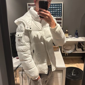 Vit Moncler pufferjacka med huva - Säljer en clean vit pufferjacka från Moncler med huva och dragkedja. Jackan har Moncler-logga på ärmen, praktiska fickor och quiltad design. Materialet är glansigt och känns lyxigt, perfekt för kalla dagar när du vill hålla stilen. Passar dig som gillar streetwear och premiumkänsla.