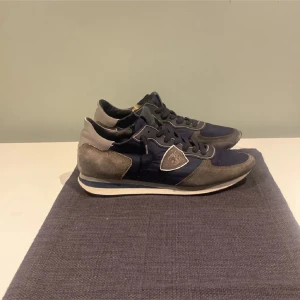 Philippe Model sneakers i blå och grå mocka - Snygga sneakers från Philippe Model i marinblått tyg och grå mocka med vita detaljer. Skorna har snörning, rund tå och en platt sula. Perfekt för dig som gillar en sportig och stilren look med premiumkänsla.