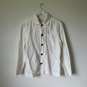 Vit overshirt från These Glory Days - Snygg vit overshirt från These Glory Days med klassisk krage, stora svarta knappar och tre fickor framtill. Tillverkad i bomull, perfekt för lager-på-lager och enkel att styla till olika outfits. Långärmad och stilren design som funkar året runt. Artnr: C369