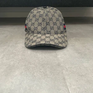 Gucci keps med GG-mönster och röd-blå detalj - Helt ny snygg keps från Gucci i beige med klassiskt GG-monogram över hela kepsen. På sidan finns en röd och blå randig detalj. Justerbar rem baktill i svart läder. Perfekt accessoar för att lyfta din outfit.