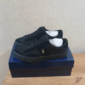 Svarta sneakers från Ralph Lauren - Stilrena svarta sneakers från Ralph Lauren, modell Sayer Sport. Skorna har låg profil, klassisk snörning och är dekorerade med den ikoniska broderade polospelaren i guld på sidan. Ovandelen är i mocka och textil för en clean look. Yttermatrialet på inne sulan(kan ras bort) har lossant lite. Men annars är de i väldigt bra  skick. Box följe med.