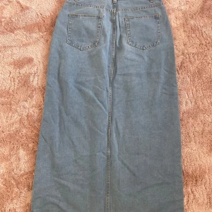 Lång jeanskjol i ljusblå denim - Säljer en klassisk lång jeanskjol i ljusblå denim med raka linjer och två bakfickor. Kjolen har bälteshällor och synliga sömmar för en tidlös look. Perfekt för dig som gillar enkel och avslappnad stil.