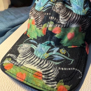 Färgglad keps med tiger- och växtmönster - Snygg keps med svart bas och färgglatt tryck av tigrar, gröna blad och röda detaljer. Kepsen har böjd skärm och är tillverkad i ett slitstarkt bomullsmaterial. Perfekt för dig som vill sticka ut med ett unikt djur- och naturmönster.