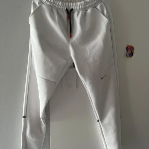 Vita mjukisbyxor från Nike, storlek M - Snygga vita mjukisbyxor från Nike med elastisk midja och svart snörning med röda detaljer. Byxorna har en clean look med diskret Nike-logga på benet och svarta ränder vid bensluten. Perfekta för chill eller träning.