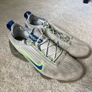 Nike Vapormax grå/blå sneakers - Nike Vapormax sneakers i ljusgrått med blå och neongula detaljer. Ovandel i stickad mesh och genomskinlig sula med synliga luftbubblor. Snygg design med snörning och blå hälflik med neongult Swoosh. Perfekta för dig som gillar sportig stil.