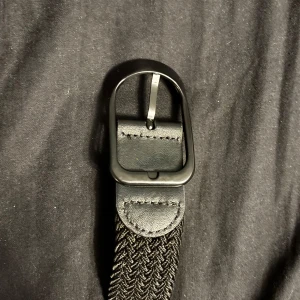 Svart flätat bälte med mattsvart spänne - Snyggt svart bälte med flätad design och mattsvart metallspänne. Bältet har detaljer i skinn vid spännet och en stilren look som passar till jeans eller kostymbyxor. Perfekt accessoar för att lyfta din outfit.