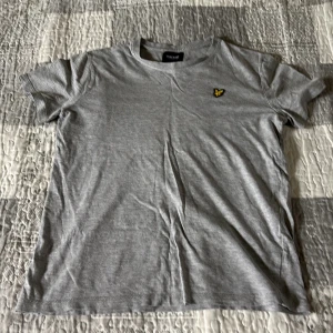 Grå t-shirt från Lyle & Scott - En stilren grå t-shirt från Lyle & Scott med klassisk passform och korta ärmar. T-shirten har en liten gul broderad logga på bröstet och är tillverkad i mjuk bomull. Perfekt för en clean och enkel look. Passar 152-158