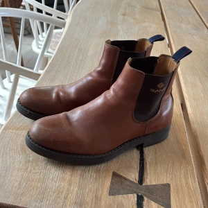 Bruna chelsea boots från Gant - Snygga bruna chelsea boots från Gant i skinn med elastiska svarta paneler på sidorna och robust svart sula. Klassisk rund tå och dragflik bak med blå och röda detaljer. Perfekta för dig som gillar stilrena och tidlösa skor.