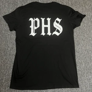 Svart PHS Support t-shirt - Svart t-shirt med stor vit PHS-tryck på ryggen och mindre tryck med knogjärn, lagerkrans och texten 'SUPPORT' på bröstet. Klassisk rund hals och korta ärmar. Skön passform och stilren streetkänsla.
