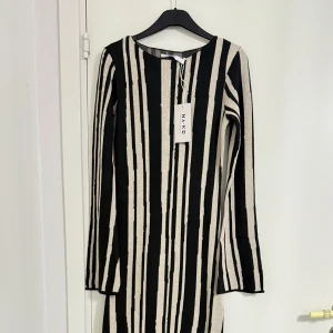 Randig långklänning från NA-KD - Snygg långärmad långklänning från NA-KD i stickad kvalitet. Klänningen har ett svart och beige randigt mönster och rak passform. Perfekt för dig som gillar en stilren och modern look med edge.