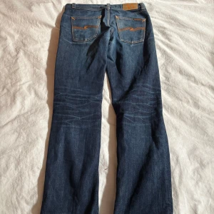 Mörkblå jeans med orange sömmar - Säljer ett par mörkblå jeans från Nudie Jeans med snygga slitningar och orange kontrastsömmar. Modellen har klassiska fem fickor och raka ben med lätt utsvängning. Jeansen har coola slitdetaljer och en autentisk tvättad look.