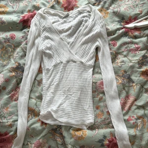 Vit omlott v-ringad ribbad tröja - Supersnygg vit ribbad tröja med omlott och djup v-ringning. Långärmad och figurnära passform, perfekt att styla med jeans eller kjol. Mjukt och stretchigt material som sitter skönt hela dagen.